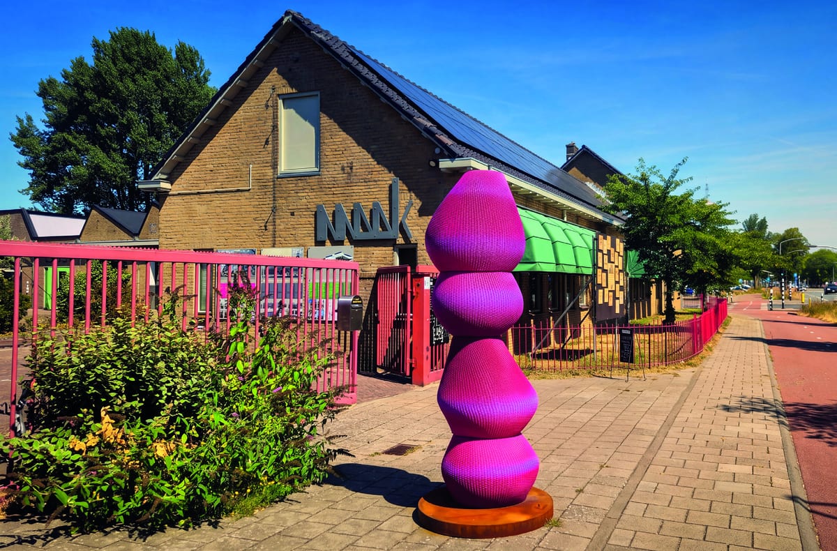 Roze gestapelde sculptuur voor de ingang van MAAK Haarlem aan de Oudeweg, met rood hek en bakstenen gebouw