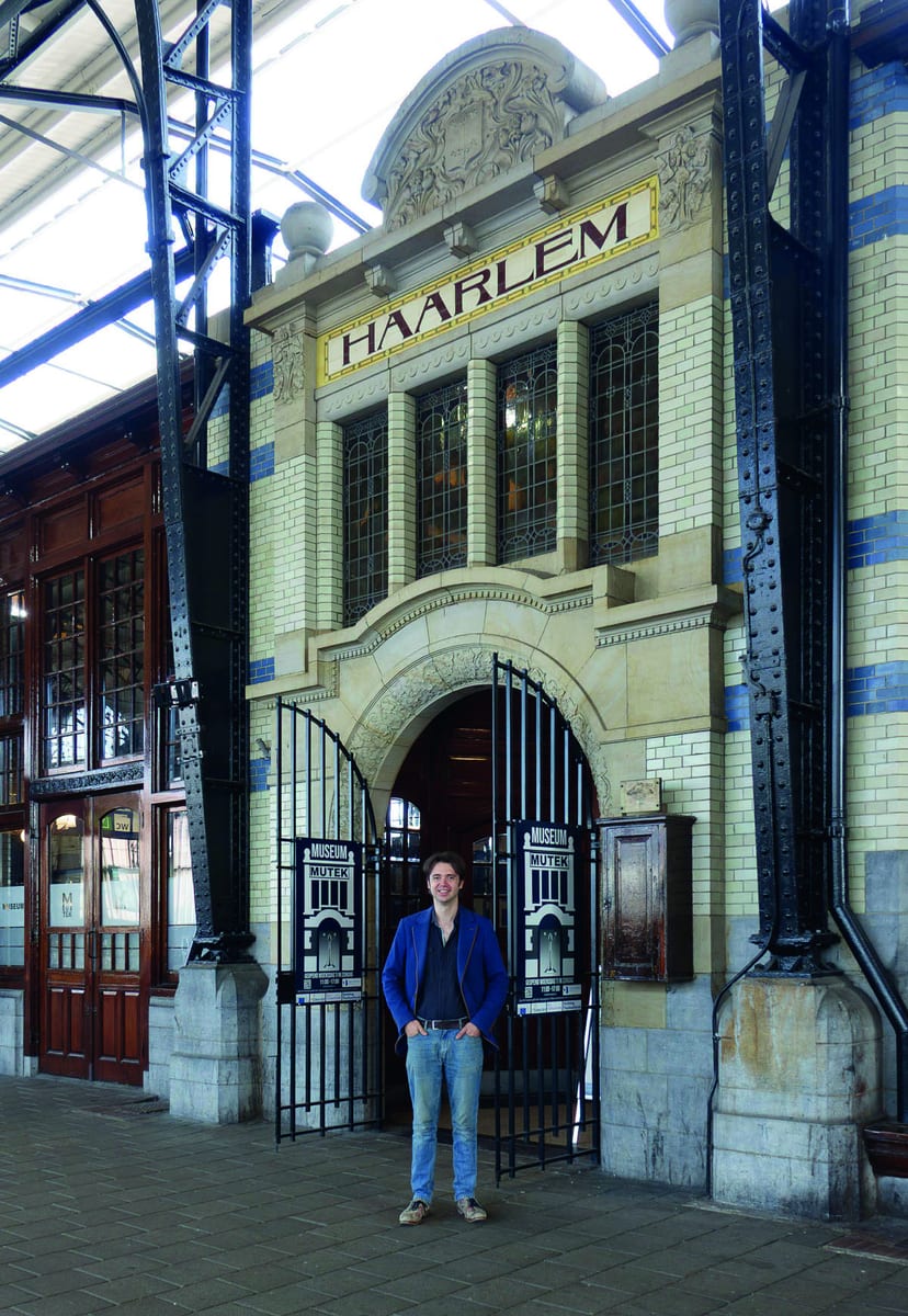 Robert van Rhijn voor de ingang van museum MUTEK op het perron van station Haarlem, onder het tegeltableau Haarlem